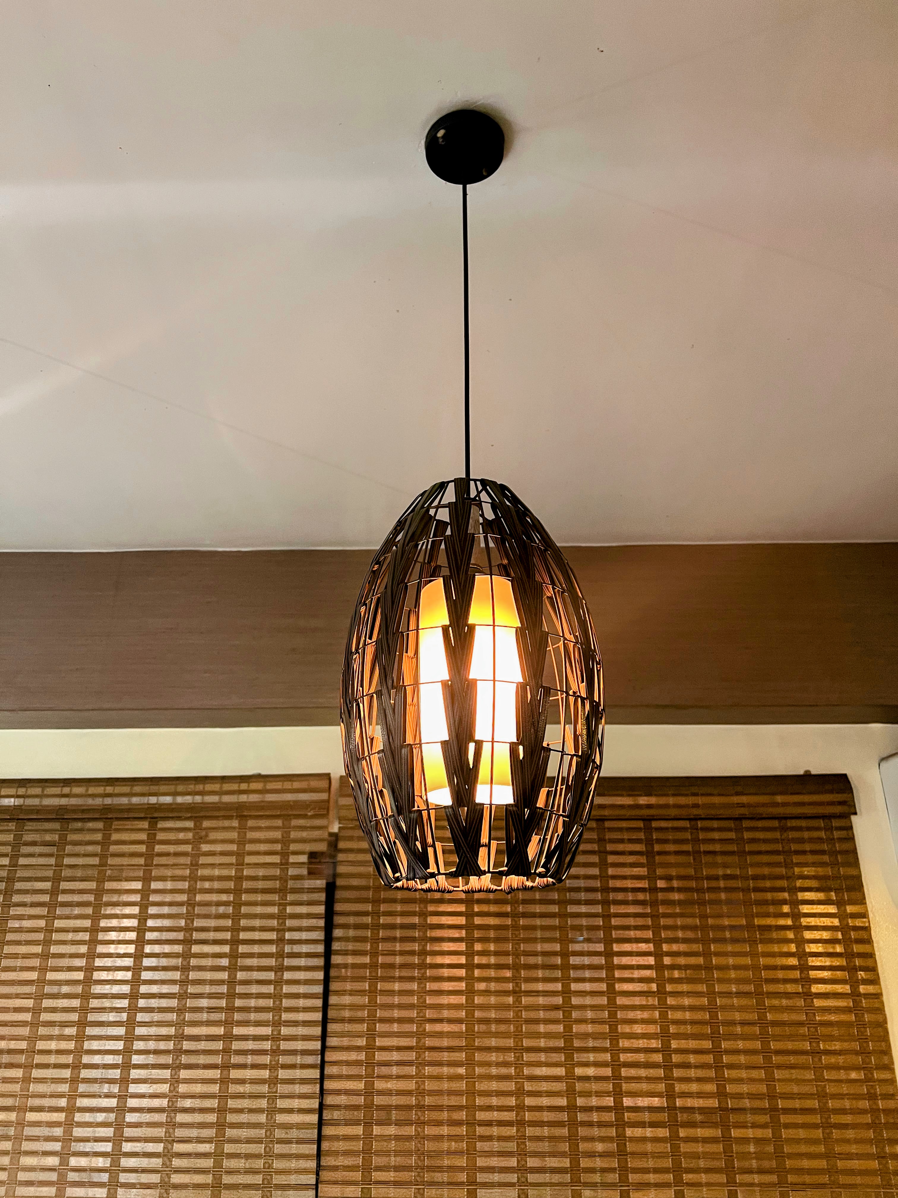 An oval woven wicker pendant light casting a warm glow in front of bamboo blinds.