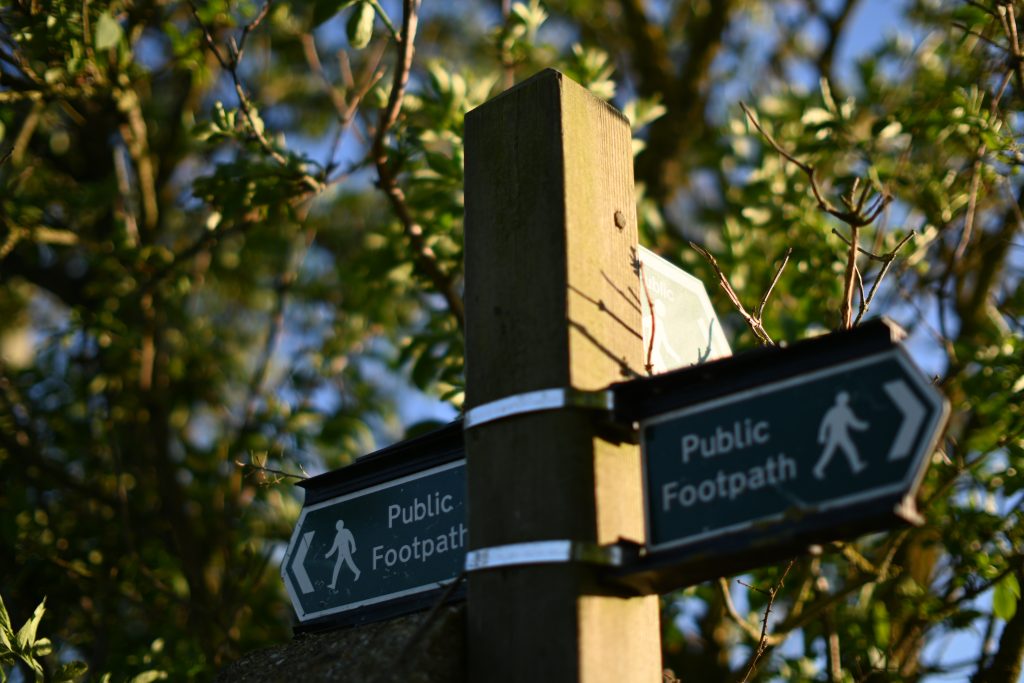 A public footpath sign in the UK, surrounded by hedgerows.