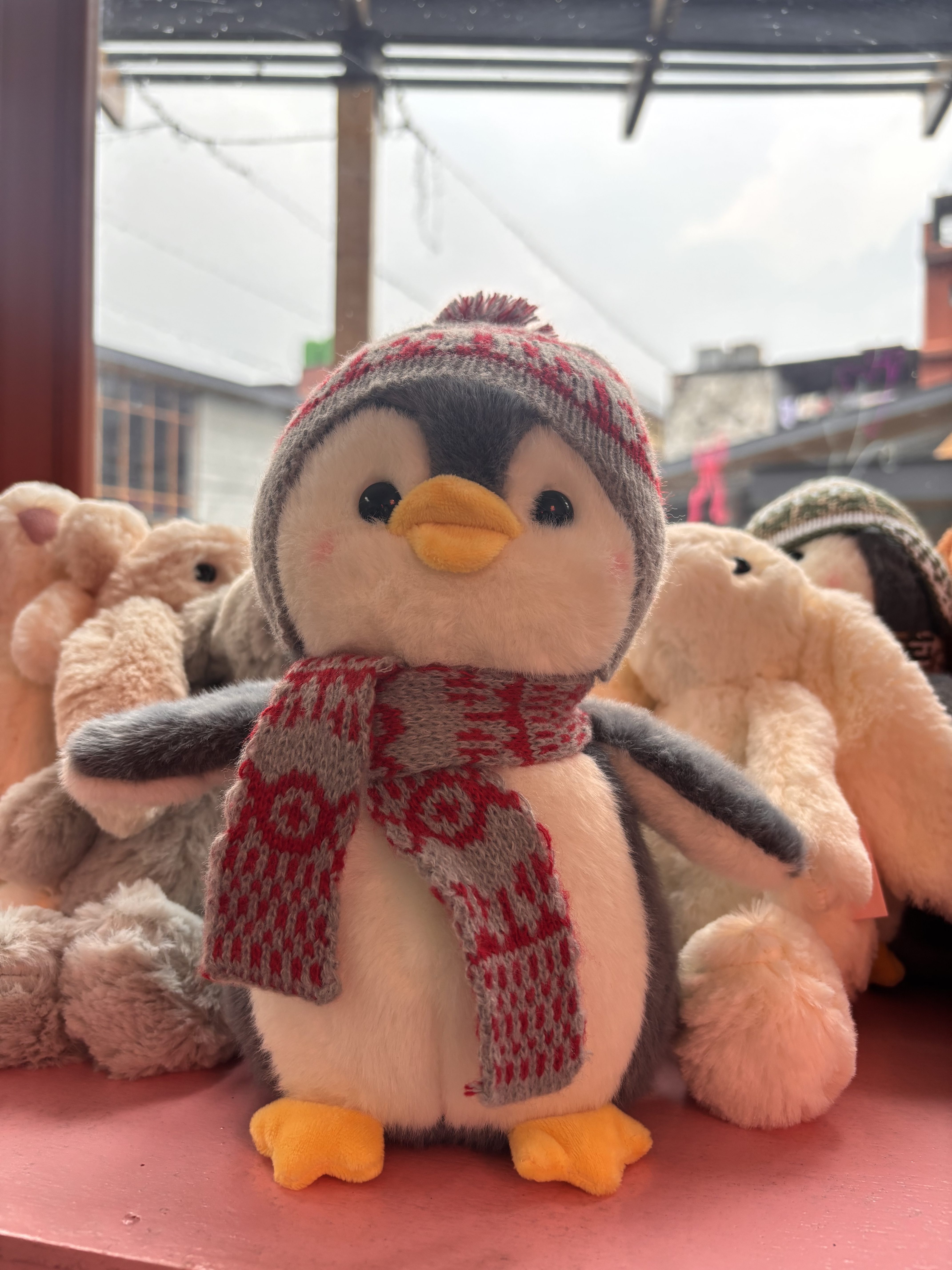 A cute penguin plushie wearing a red and gray knitted hat and scarf, surrounded by fluffy bunny toys.