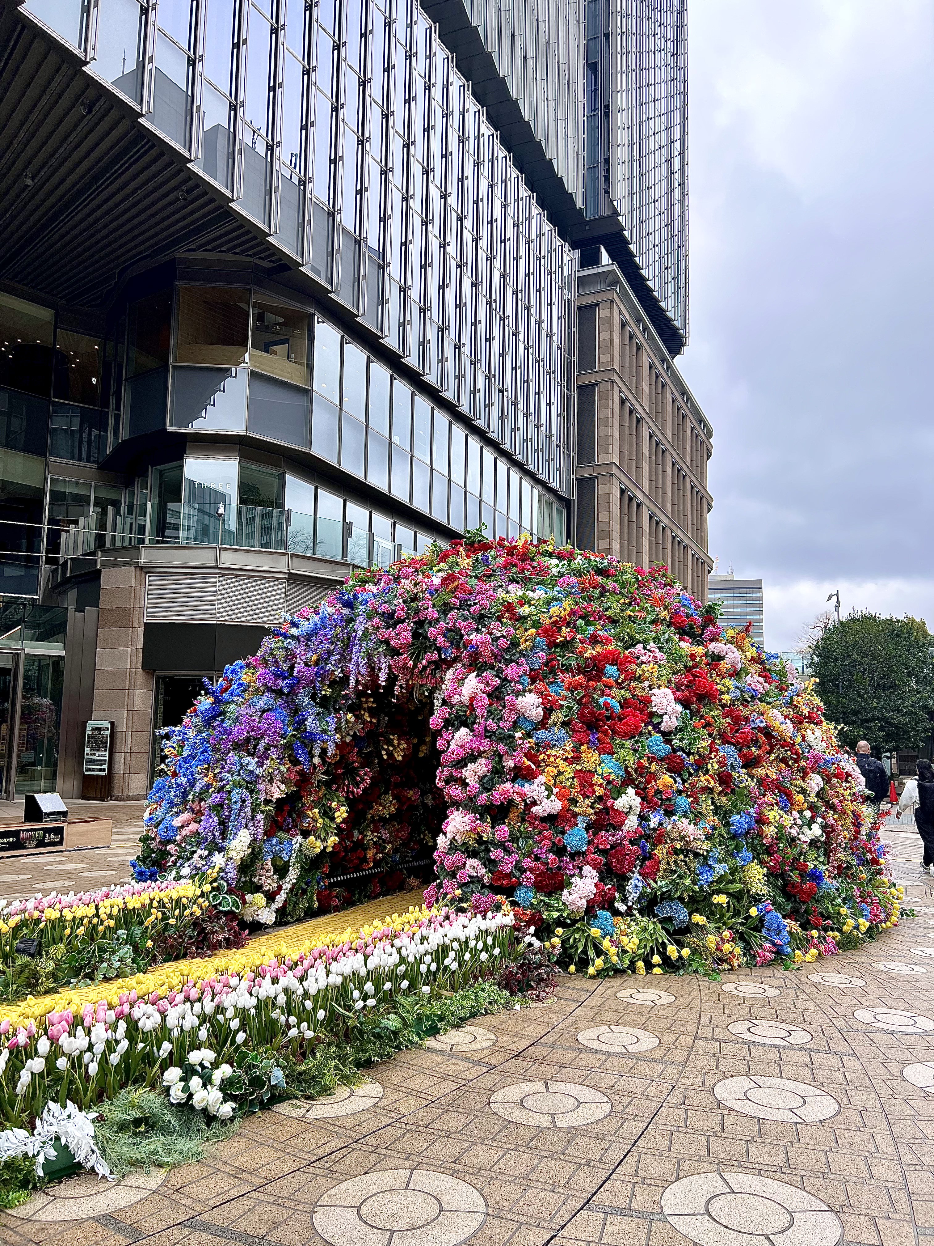A large dome-shaped flower installation covered in colorful blooms, set in a city plaza in front of tall modern buildings.