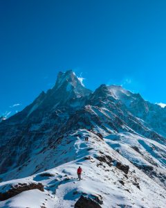 Snow-covered mountain ridge with hikers trekking toward a dramatic Himalayan peak under a deep blue sky.
