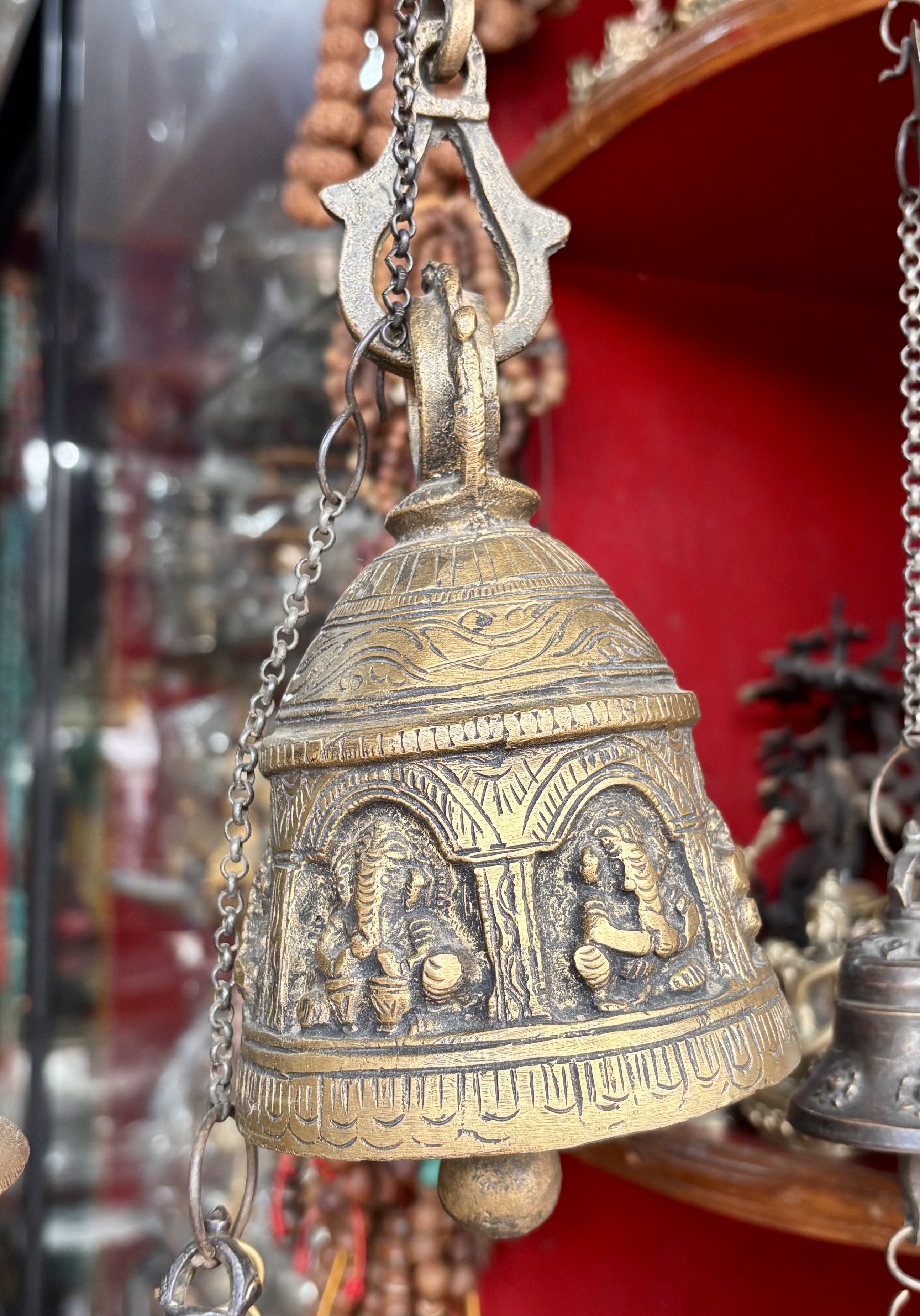 A close-up of a brass temple bell with intricate carvings, hanging by a chain in front of a red background and other decorative items.