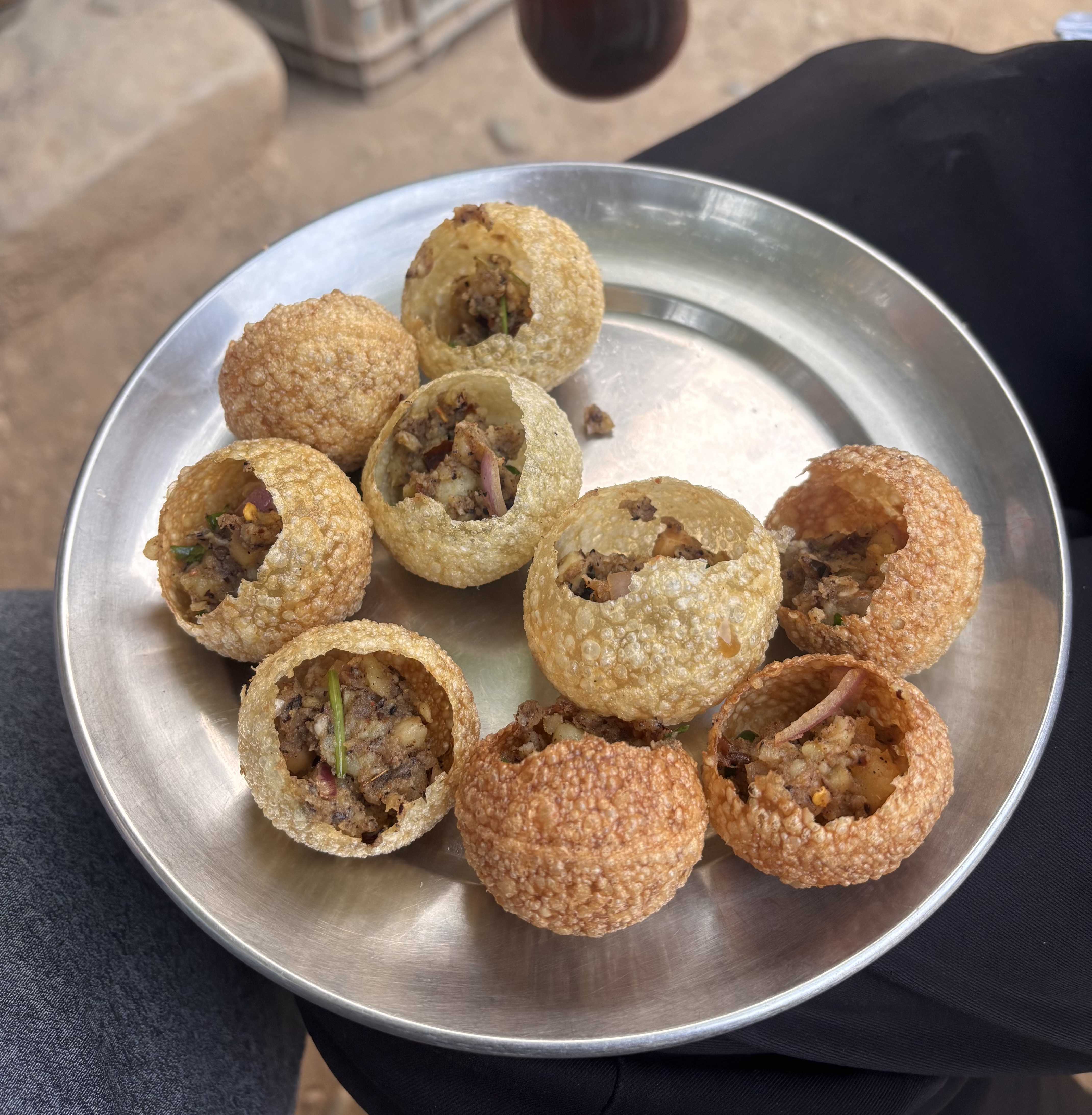A stainless steel plate holds several hollow, crispy spheres filled with a mixture of spices, vegetables, and meat. The spheres, commonly known as golgappas or panipuri, vary in color and texture, with some being golden brown and others lighter in hue.