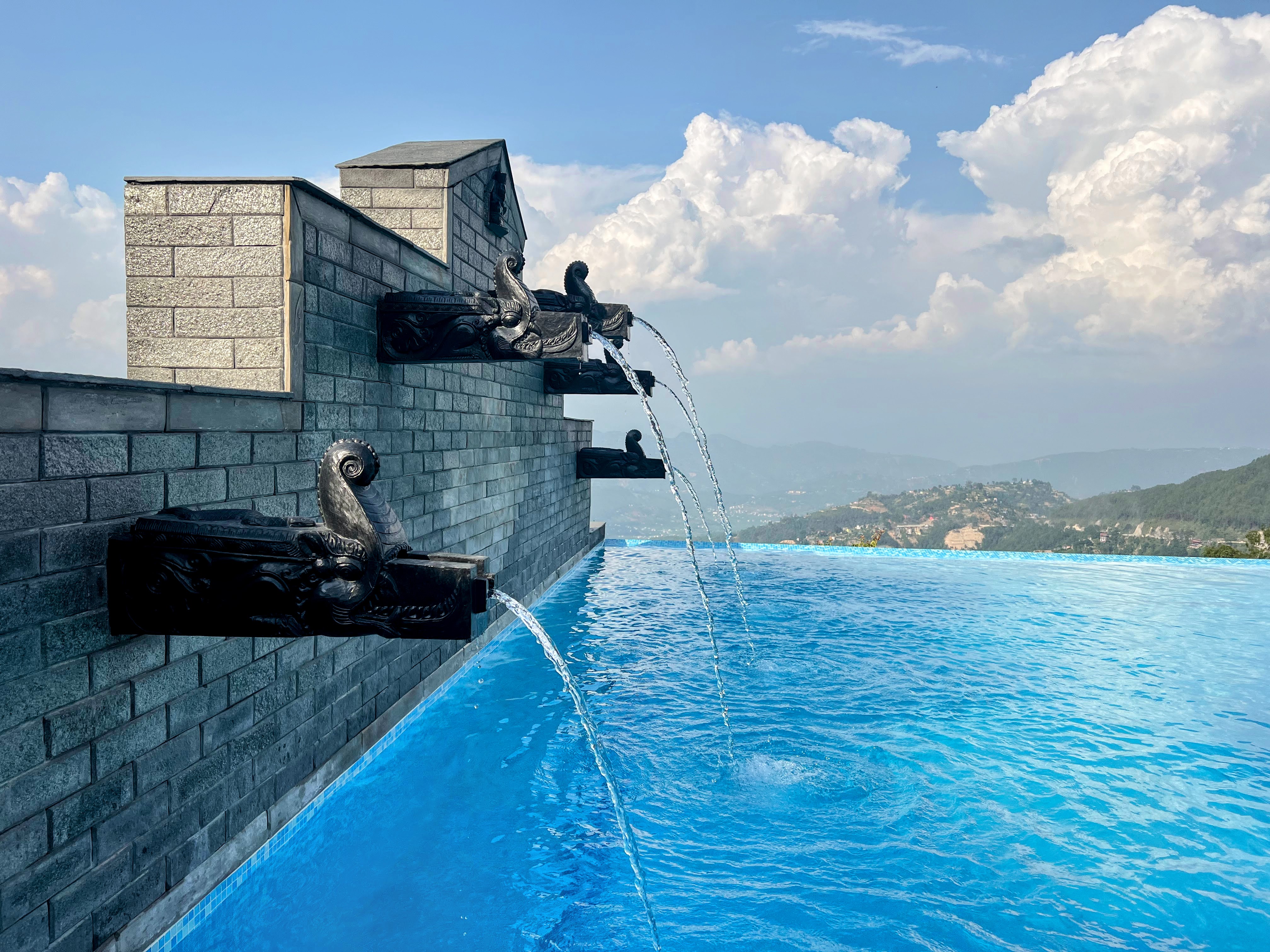 Traditional black stone spouts pouring water into a bright blue infinity pool with mountains in the background.