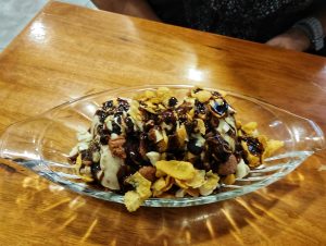 A glass boat-shaped dish filled with ice cream topped with a variety of crunchy cereal pieces, drizzled with chocolate syrup.