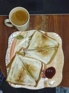 Toasted sandwiches on a floral plate, accompanied by a dollop of ketchup. A cup of tea sits beside on a wooden table, creating a cozy breakfast vibe.