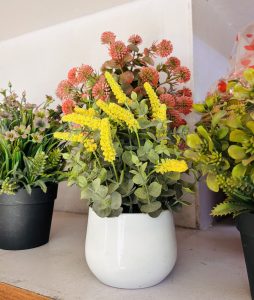 A white ceramic pot holds vibrant yellow flowers with lush green leaves, surrounded by artificial plants in pink and green tones.