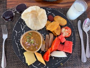 A top-down view of a breakfast plate featuring fried flatbread, a bowl of chickpea curry, sausages, potato wedges, fresh fruit slices, and a glass of milk. 