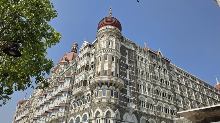 A low-angle shot of the iconic Taj Mahal Palace Hotel in Mumbai, showcasing its grey stone facade, intricate white arched balconies, and prominent red-tiled domes against a clear blue sky.