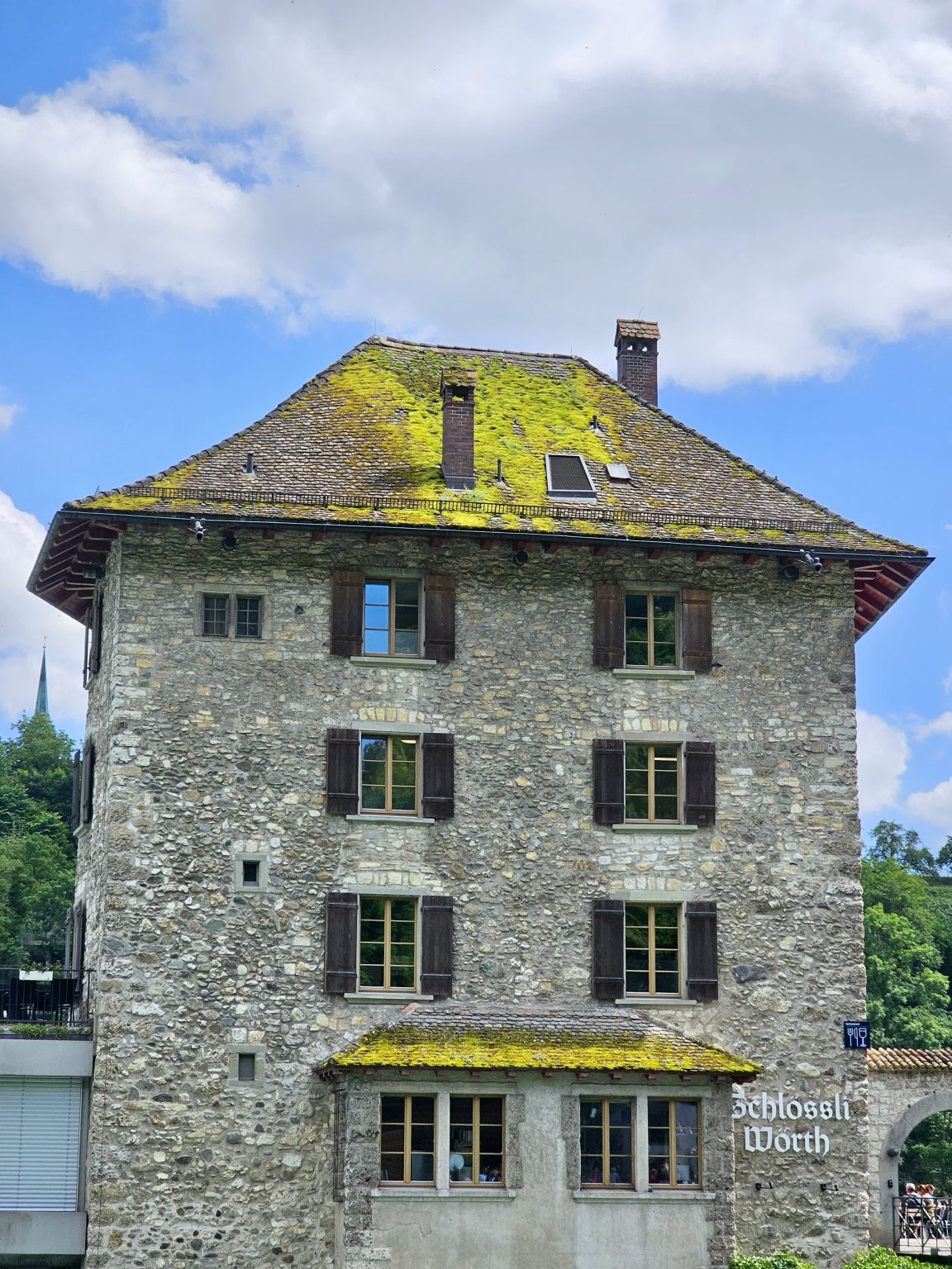 An old stone building, Schlössli Wörth (also known as Wörth Castle), a 13th century castle with wooden windows and a moss-covered roof, stands under bright clouds and blue skies near the Rhine Falls in Switzerland. Its peaceful architecture reflects a classic Swiss charm.