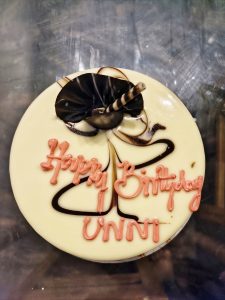 White birthday cake with chocolate decorations and pink “Happy Birthday UNNI” icing.