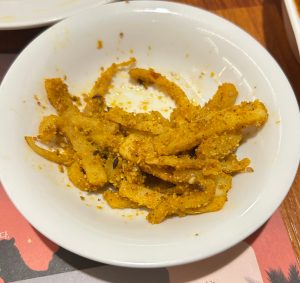 A white bowl filled with crispy golden-brown seasoned pickles sits on a wooden table.