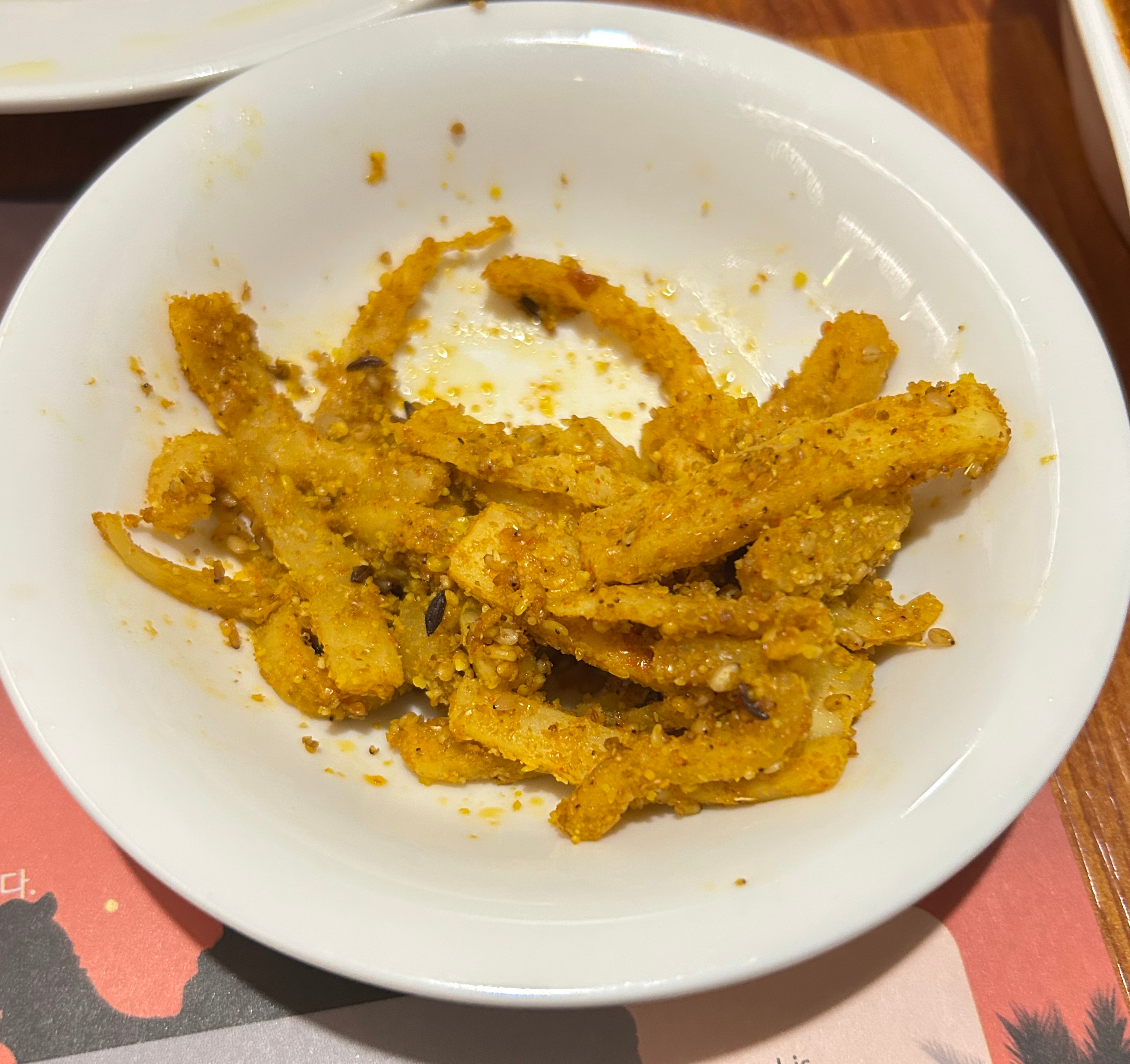 A white bowl filled with crispy golden-brown seasoned pickles sits on a wooden table.