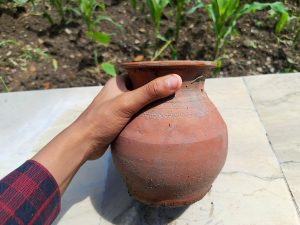 A hand holding a small traditional clay pot outdoors on a sunny day. The brown earthen pot has a rounded shape and rustic texture, with soil and green plants visible in the background.