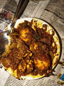 A close-up of a spicy dish with eggs, meat, and vegetables in a dark brown sauce, served in a bowl on newspaper with a metal spoon.