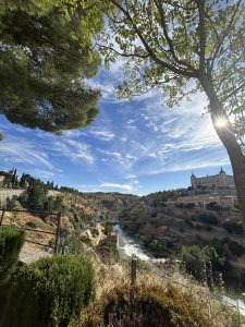 A panoramic view of a landscape featuring a winding river surrounded by rocky terrain and scattered vegetation. To the right, a large, distinguished building with turrets sits atop a hill, partially shaded by nearby trees. The sky is bright blue with wispy clouds, creating a sense of openness and tranquility. 