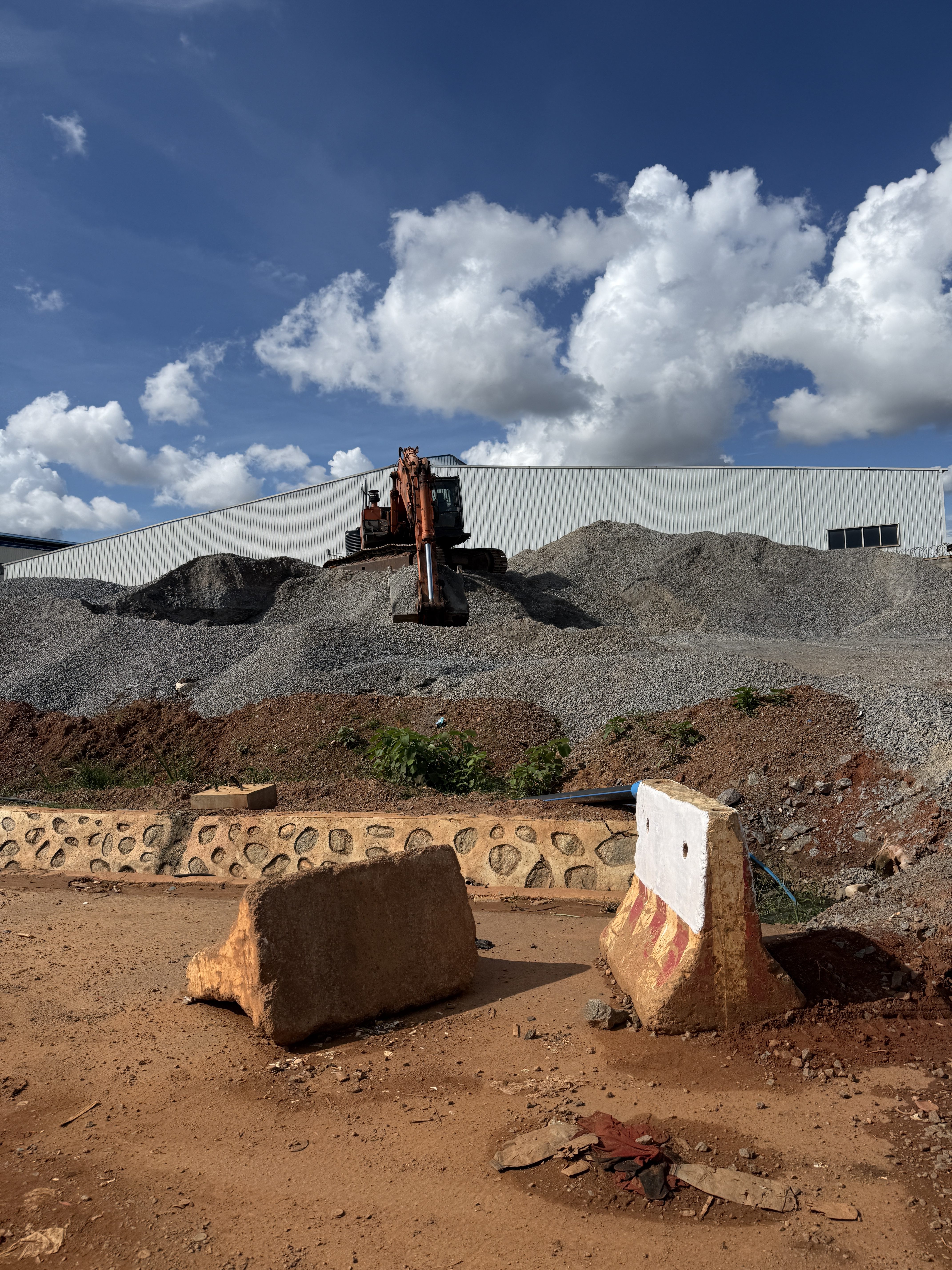 A construction site features a digger on a large gravel mound beneath a bright blue sky with fluffy clouds.