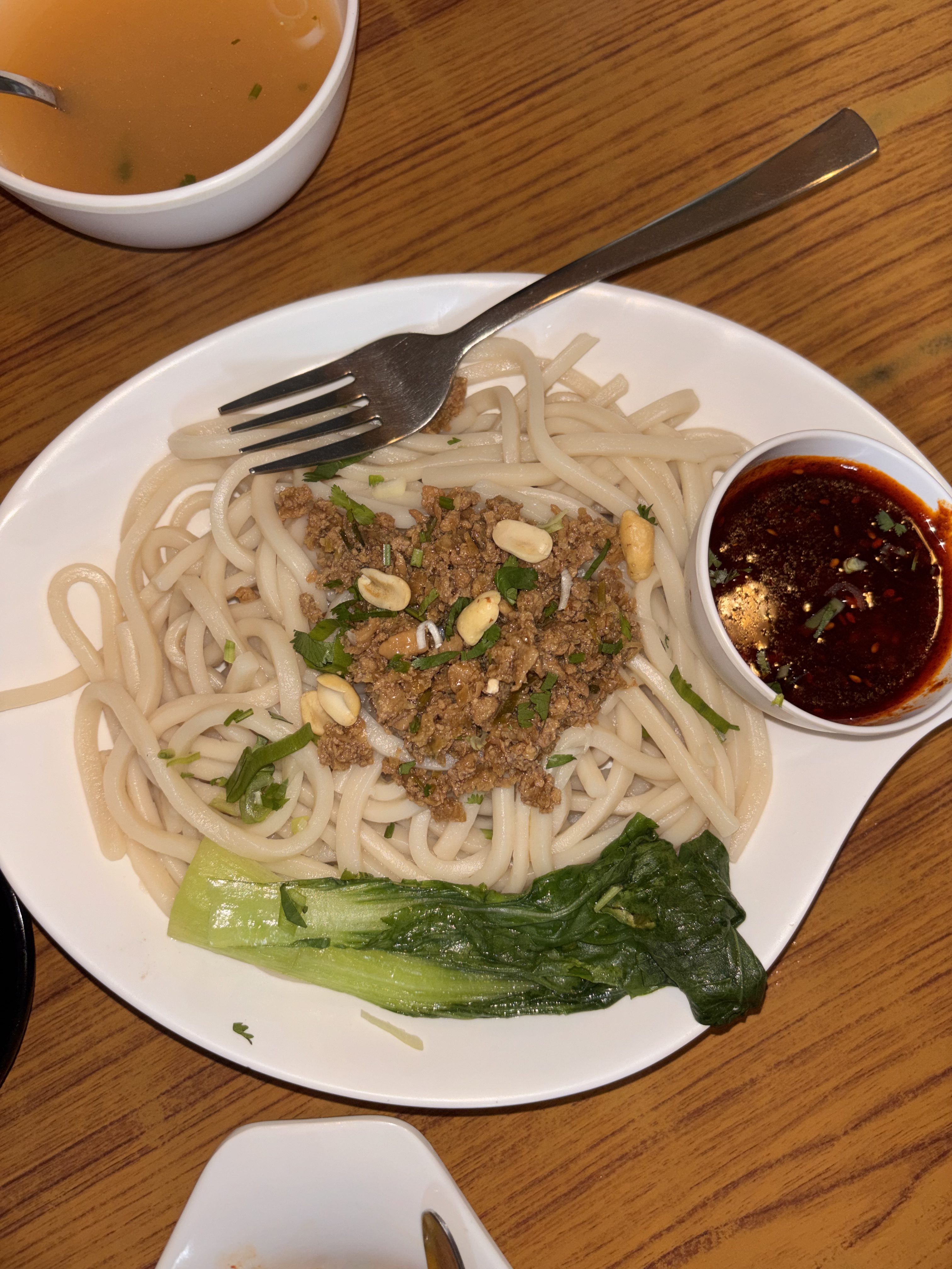 A white plate with thick noodles, topped with minced meat, peanuts, and cilantro, served with bok choy and a side of red sauce