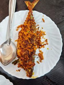 The full body of a fried mackeral, coated in breading and spices, on a white plate with a spoon.