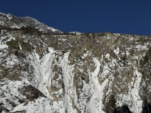 Snow-dusted rocky mountain slopes under a clear blue sky. Sparse trees cling to the rugged terrain, creating a sense of tranquility and natural beauty.