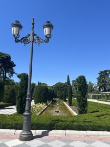 An elegant wrought-iron streetlight with two ornate lanterns stands beside a formal garden featuring trimmed hedges, a rectangular pool with small fountains, and a clear blue sky.