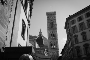 Black and white view of Florence Italy's Duomo cathedral and Giotto's bell tower framed between narrow historic buildings on a city street.