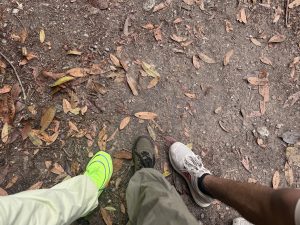 Three people stand on a dirt path covered with dry leaves. Visible are bright green, brown, and white sneakers.