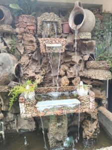A decorative stone and ceramic fountain lets water flow through multiple levels, creating a relaxing and handcrafted scene surrounded by rocks, plants, and rustic clay pots.