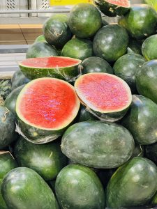 A stack of shiny green watermelons, with a few cut open showing bright red flesh inside.