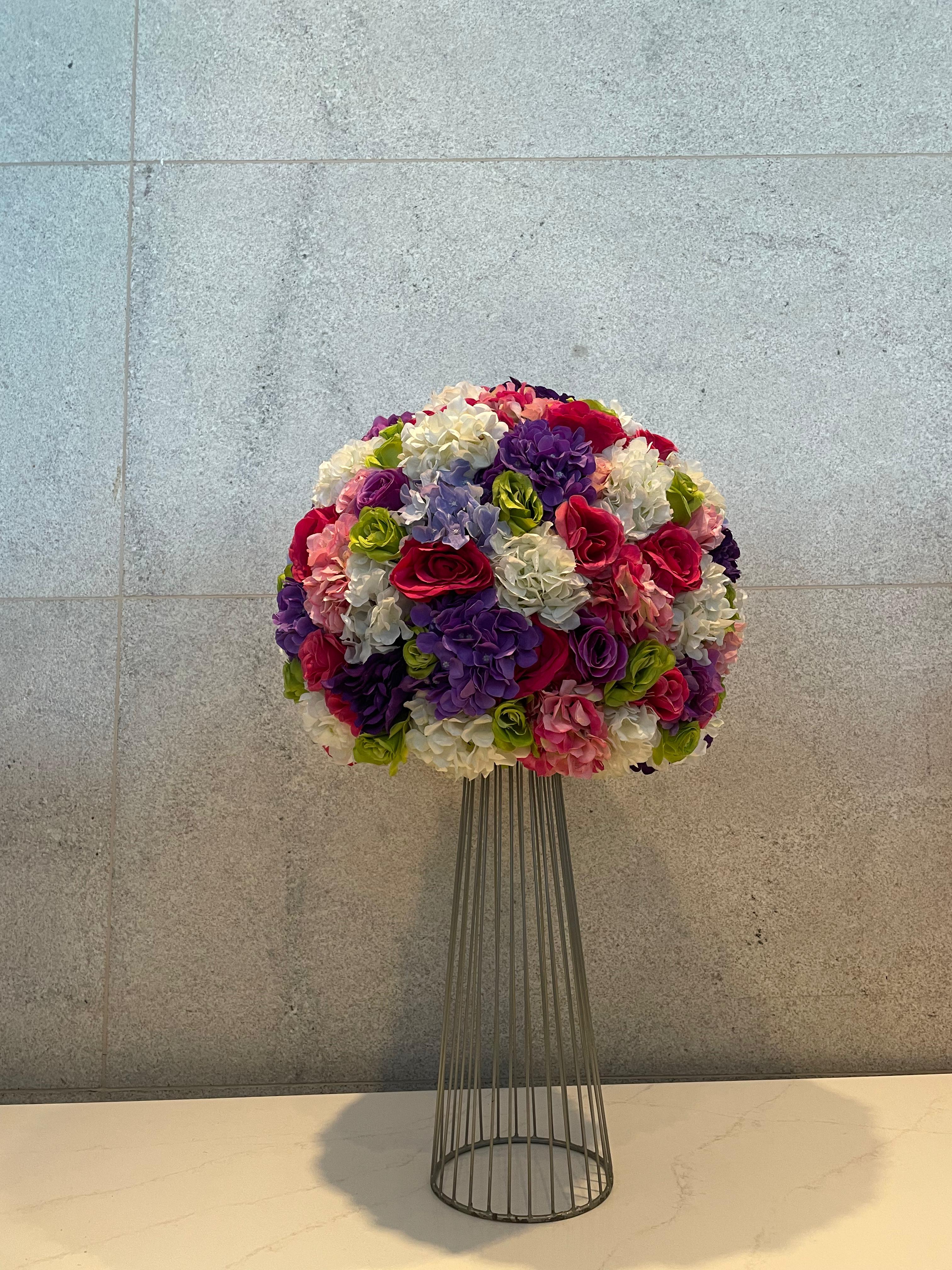 A vibrant, spherical floral arrangement featuring pink, purple, white, and green flowers, resting on a tall, slender metal wire stand against a plain, textured grey wall.