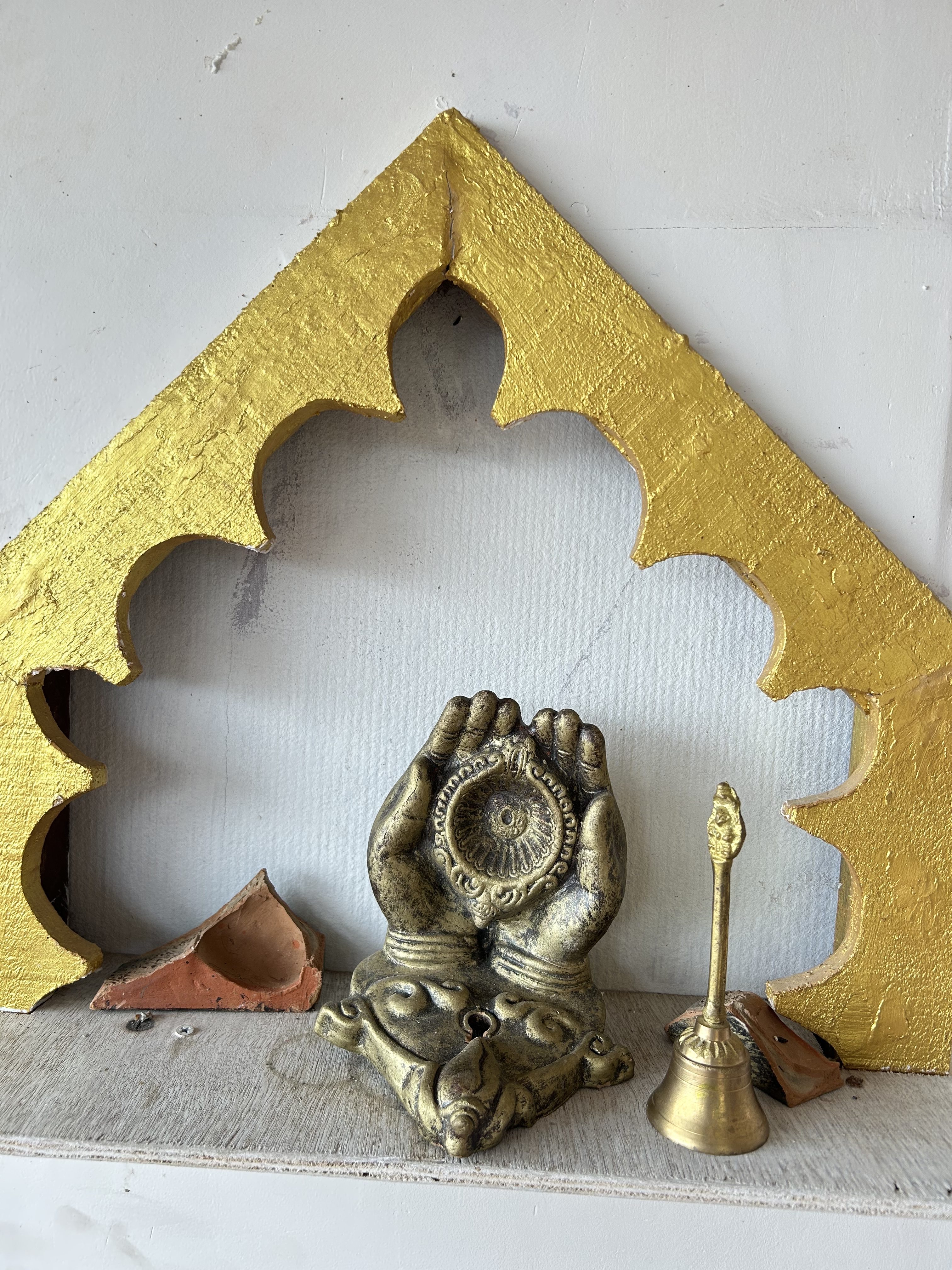 Golden decorative arch frames a brass sculpture of two hands holding a circular design, flanked by a brass bell and terracotta pieces.