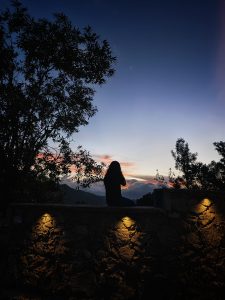 This is an evening scene, so it's mostly dark. In the foreground is a stone wall with some ambient lights pointing down. A person with long hair is sitting on the wall facing away from the camera. They're seen only in silhouette. There are trees around them. In the distance are some hills with clouds lit by the sun from eblow the horizon.