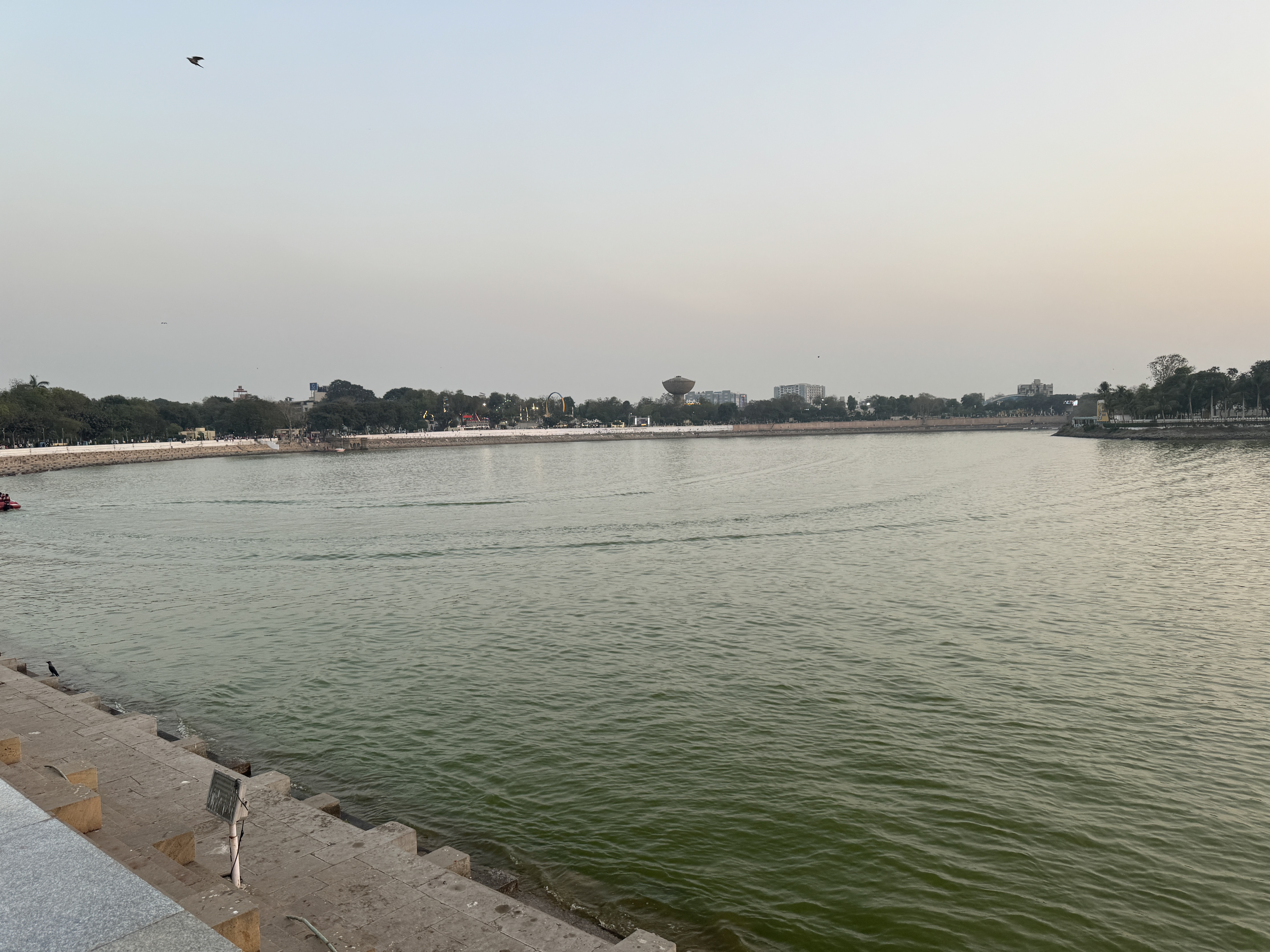 A wide view of a calm, large lake with stone steps along the foreground shoreline and a distant city skyline under a pale sky.