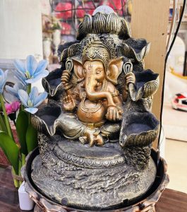 Bronze Ganesh statue with intricate detailing, seated in a meditative pose on a decorative base, surrounded by blue flowers and small trinkets.