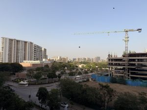 A wide urban cityscape featuring residential and commercial buildings under a clear sky. A construction crane stands beside an unfinished structure, while trees and roads add depth to the scene.