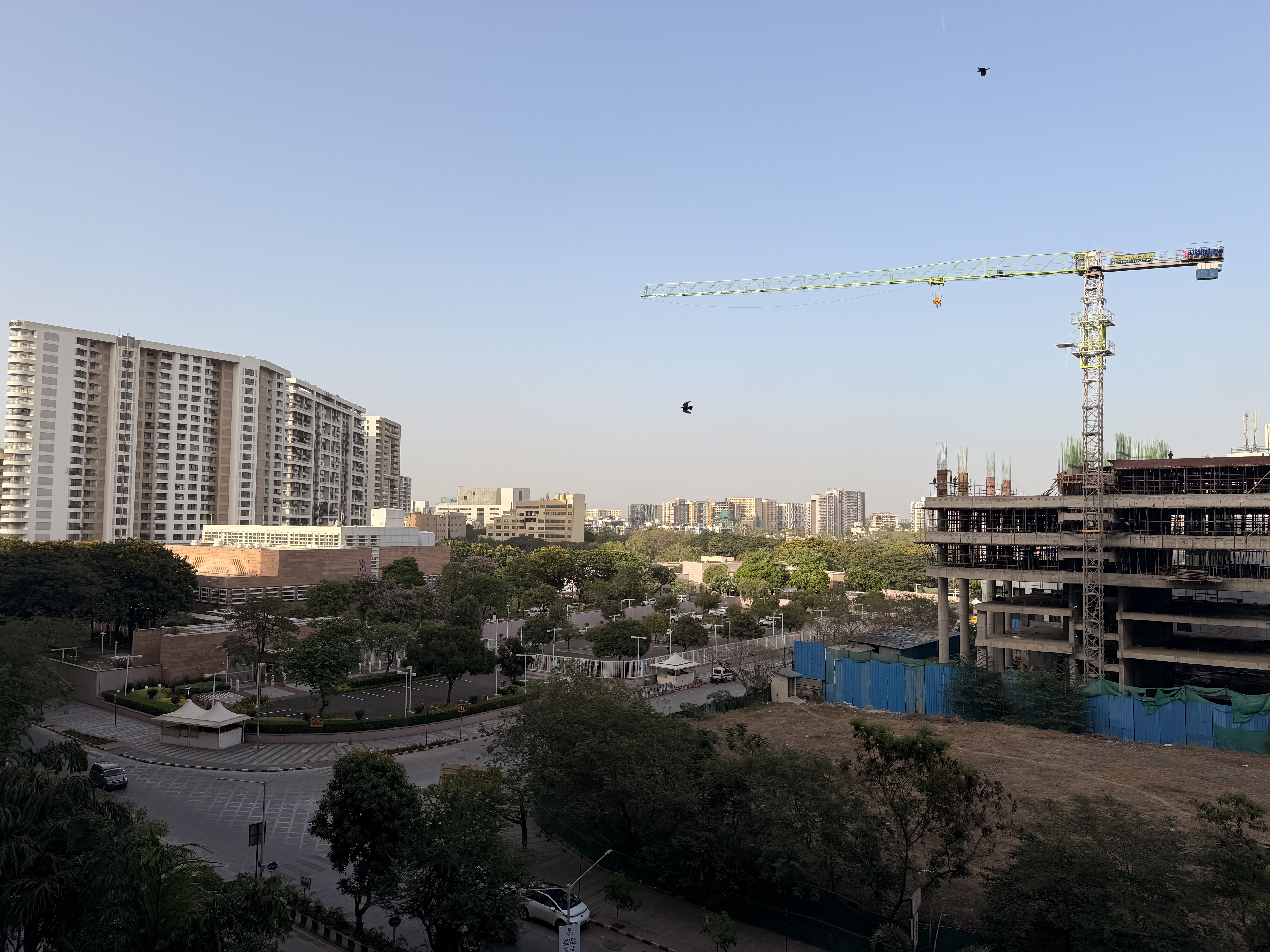 A wide urban cityscape featuring residential and commercial buildings under a clear sky. A construction crane stands beside an unfinished structure, while trees and roads add depth to the scene.