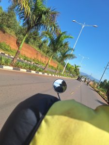 A view from a motorcycle showing the handlebars and the rider’s yellow shirt, with a straight road lined by palm trees and shrubs leading toward distant vehicles under a clear blue sky in Kigali