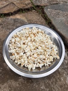 A metal bowl filled with freshly popped popcorn sits on a stone surface outdoors.