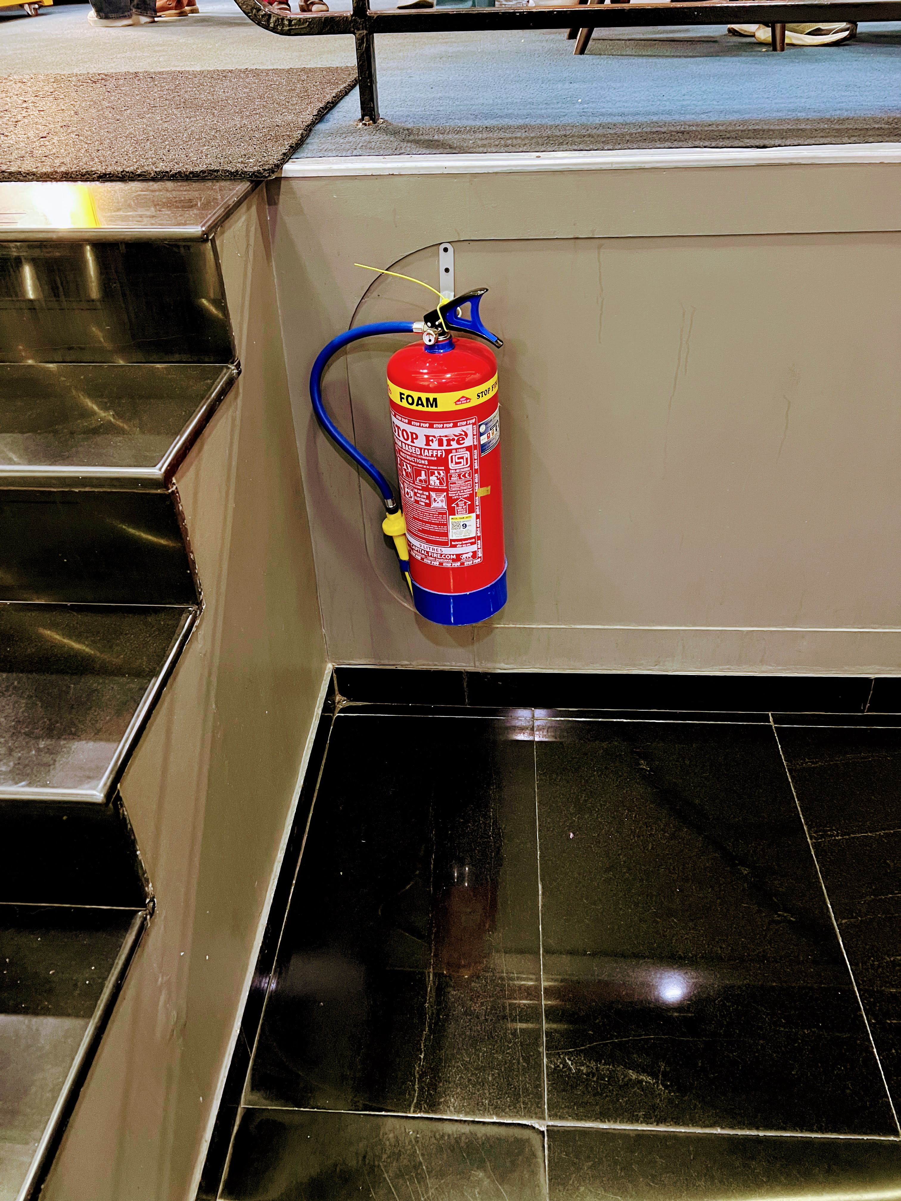 A red and blue foam fire extinguisher mounted on a wall next to black tiled stairs.