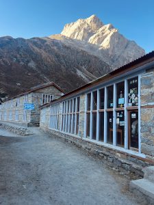 Stone buildings with large windows line a dirt path against rugged sunlit mountains, conveying a tranquil atmosphere.