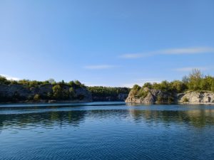 A sunny view of a flooded limestone quarry with deep blue water, green-covered cliffs, and a clear sky.