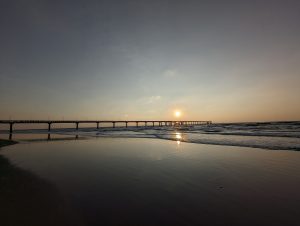 A long bridge known as Narayani Bridge of Nepal, stretching across a beach and into the ocean during a hazy sunset, with the sun reflecting on the wet sand and gentle waves