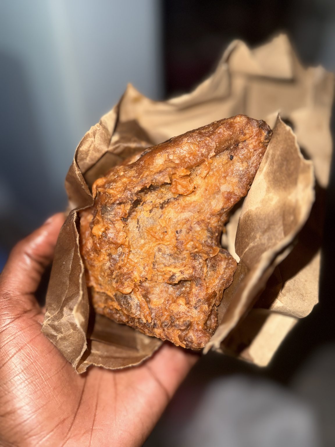 A piece of fried chicken wrapped in a paper held in a hand.