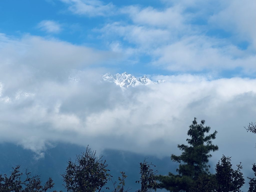 A serene mountain scene with snow-capped peaks partially obscured by fluffy clouds. Foreground trees add depth; the sky is a bright blue.
