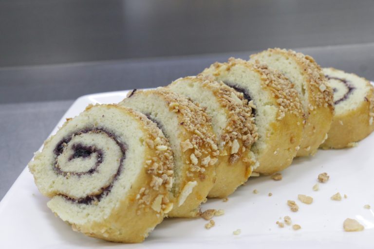 Slices of a jelly roll swiss cake with crushed nuts on top are arranged on a white plate. The cake is spiraled with a dark filling, creating a tempting dessert display.
