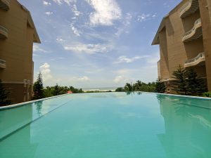 Infinity pool with clear blue water stretching towards the ocean horizon, flanked by two brown buildings and lush greenery under a partly cloudy sky.
