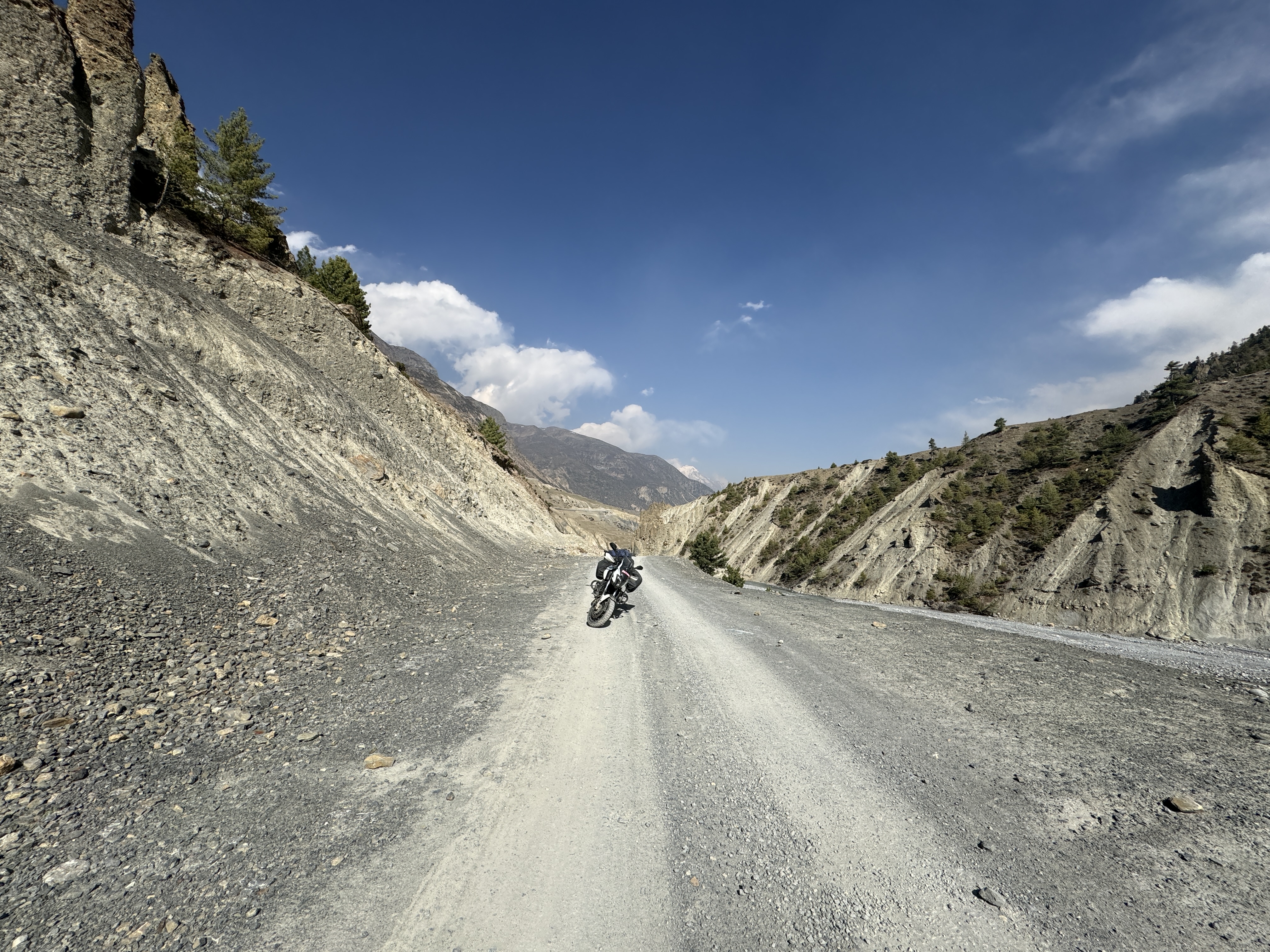A black and white adventure motorcycle with luggage is parked in the center of a wide, unpaved gravel road. The road runs through a deep, arid valley flanked by steep, eroded grey scree cliffs and sparse green pine trees under a bright blue sky with scattered white clouds in Manang, Nepal.