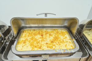 A stainless steel serving tray filled with a creamy yellow dish, topped with golden crumbs and surrounded by other trays with different food items. 
