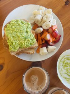 An image of a breakfast plate with toast and avocado paste on its top, besides it is a different fruits including banana, carrot and peach placed. A cup of tea is visible on the side of the plate.
