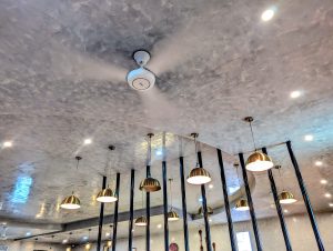 The image shows a modern interior ceiling featuring a shiny, textured surface with a subtle gray hue. There is a ceiling fan near the center, and several pendant lights with round, golden shades hanging from the ceiling.
